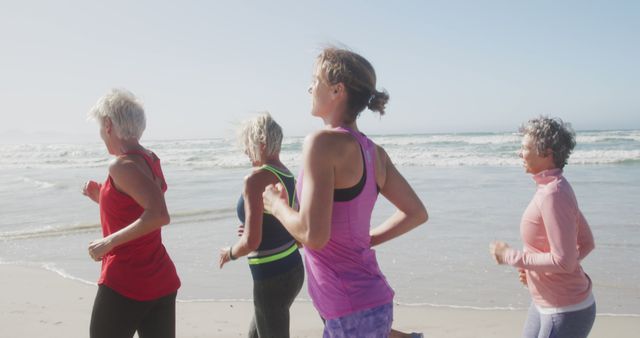 Group of women jogging on beach near ocean waves under clear sky. Fit older women embracing healthy lifestyle and physical fitness. Useful for promoting exercise, wellness programs, senior activities, beach fitness events, and outdoor group exercises.