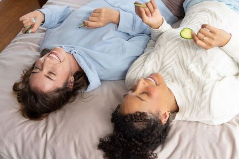 Multicultural friends laughing and relaxing on bed enjoying cucumber skincare pamper