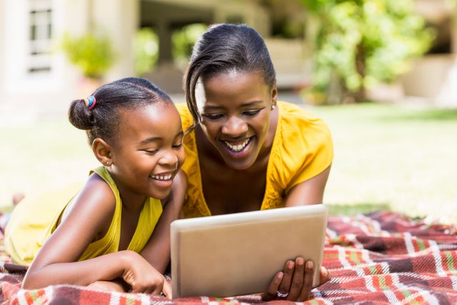 African American mother and daughter lying on a blanket in a park, smiling while using a tablet. Perfect for themes related to family bonding, technology use, outdoor activities, and happy moments. Ideal for advertisements, blog posts, and social media content promoting family time, digital devices, and outdoor leisure.