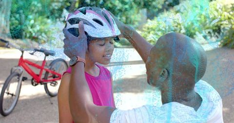 Parent securing bike helmet on child in park with red bicycle and leafy background