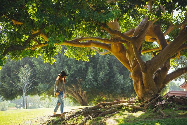 The photo shows a young woman strolling under a large, spreading tree in a sunny park. She is dressed casually and appears to be enjoying the peaceful environment. This image can be used for websites and articles related to relaxation, outdoors, nature walks, and stress relief.