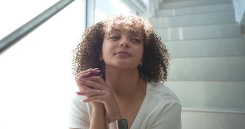 Contemplative african american woman sitting on modern staircase clasping hands in sunlight