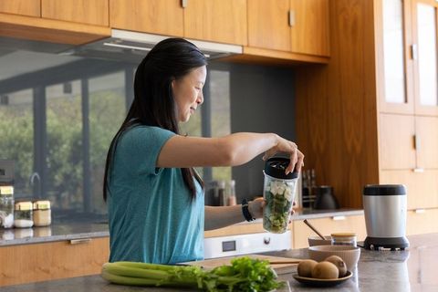 Mature asian woman preparing green smoothie in modern kitchen tightening blender lid