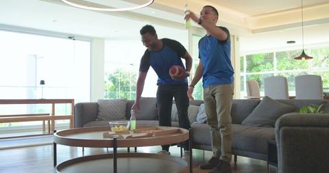Diverse men cheering and raising bottles while holding football in modern living room