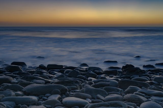 Rocky beach with smooth pebbles and gentle waves under a colorful sunset sky. Suitable for travel blogs, nature websites, meditation and relaxation content, coastal tourism promotions, and screen savers.