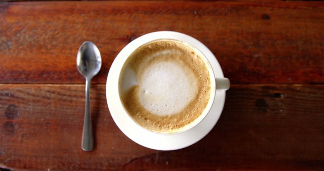 High-angle view of cappuccino cup with frothy milk placed on rustic wooden table. Metal spoon laying next to the cup. This image can be used for coffee shop promotions, cafes, beverage advertisements, breakfast menus, lifestyle blogs, or food and drink-related articles.