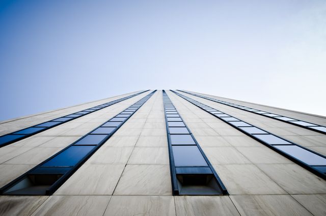 Capturing a unique perspective, this image of a modern office skyscraper framed against a clear blue sky highlights urban architecture. Excellent for use in corporate presentations, business websites, architecture portfolios, real estate industry marketing, and blogs about city life or architectural design.
