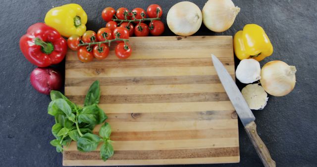 Various colorful fresh vegetables like bell peppers, tomatoes, onions, garlic, basil are arranged around a wooden cutting board with a kitchen knife. This is perfect for cooking blogs, healthy eating advertisements, cooking tutorials, and recipe websites.