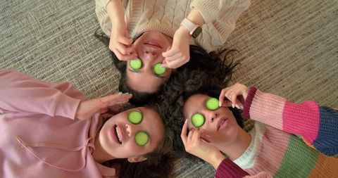 Multiracial friends lying overhead with cucumber slices on eyes relaxing in cozy knit sweaters