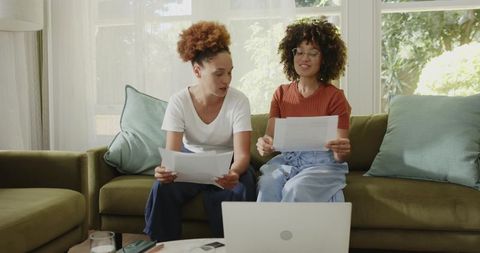 Diverse mid-adult women reviewing documents on couch with laptop in bright living room