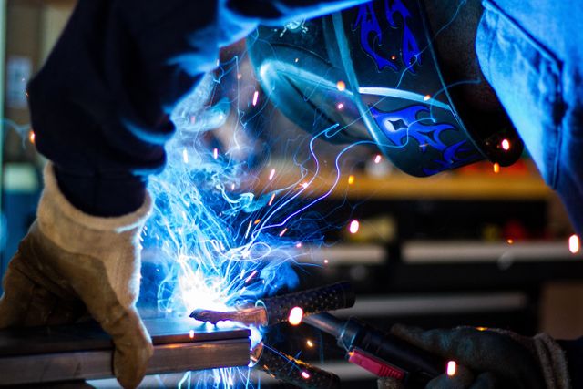Close-up of a welder engaging in metalwork with bright sparks and blue light. Welder is wearing safety gear including gloves and a protective mask. Ideal for use in articles about industrial work, metal fabrication, safety procedures, skilled trades, and occupational labor.