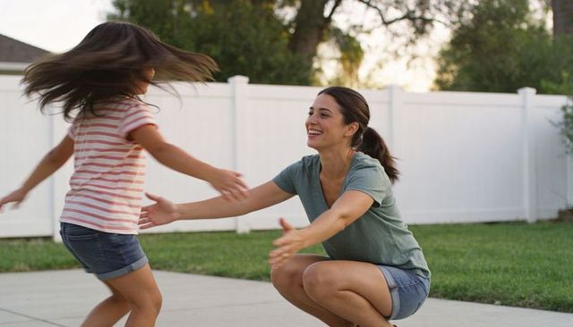 Joyful mother crouching with arms open welcoming daughter running on suburban backyard patio