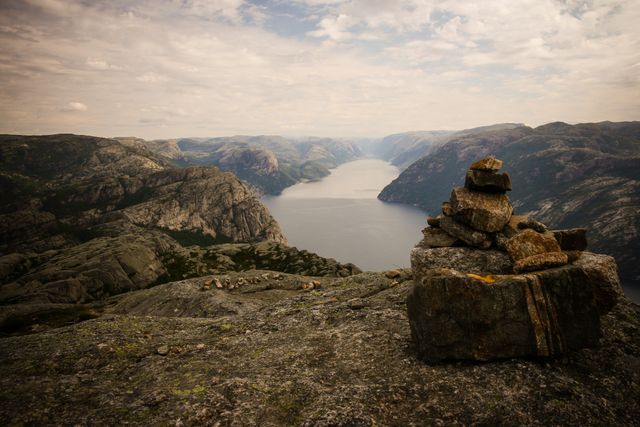 Captures a stone cairn positioned on a rocky outcrop with a stunning view of a fjord surrounded by mountains. This setting evokes a sense of peaceful wilderness and is ideal for use in travel brochures, outdoor adventure promotions, and nature-themed publications.