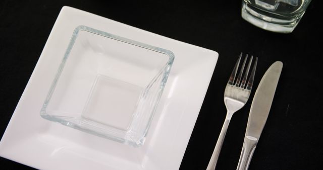 A square white plate is paired with a smaller square glass dish, accompanied by a fork and knife on a dark surface, with copy space. The arrangement suggests a modern and minimalist table setting, ready for a meal.