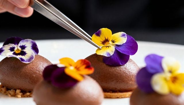 Pastry chef placing edible pansy on chocolate mousse dome using tweezers gourmet plating