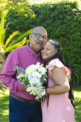African american senior couple embracing in sunny garden holding white bouquet, joyful