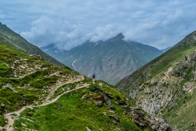 Solo hiker navigating winding trail in lush green mountains on cloudy day with majestic peaks. Ideal for promoting outdoor adventure travel, hiking gear, ecotourism, nature retreats, and environmental campaigns.