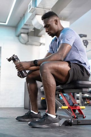 African American man resting on bench holding shaker bottle after workout at gym