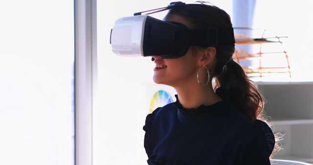 Woman in casual attire using virtual reality headset indoors, illustrating the use of modern technology in daily life. Can be used to highlight advancements in VR tech, digital experiences, gaming, and immersive environments. Suitable for tech blogs, virtual reality updates, educational content about digital innovation, and promotional materials for VR products.
