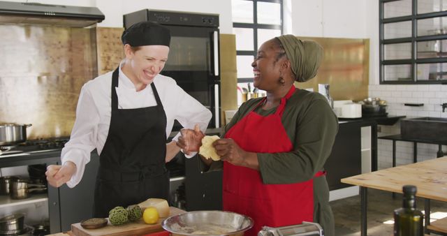 In this vibrant culinary scene, two female chefs are seen collaborating and enjoying their time cooking together in a commercial kitchen. One is dressed in traditional chef attire, and the other, more casually dressed with a headscarf, share a moment of laughter. Fresh ingredients and kitchen equipment are seen on the counter, indicating active food preparation. This image is ideal for demonstrating culinary teamwork, diversity in the kitchen, and can be used in content related to cooking classes, chef training programs, or restaurant advertisement.