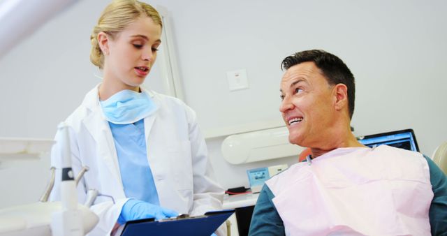 Young female dentist is discussing a treatment plan with a male patient seated in a dental chair in a modern dental clinic. The patient looks engaged while the dentist, holding a clipboard, explains the procedure. Useful for illustrating content focused on dental health, professional care, patient communication, and healthcare advertisements.