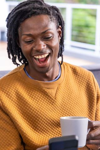 African american man laughing while checking smartphone and holding coffee on balcony sofa