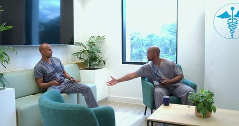 African american healthcare workers relaxing and greeting in clinic breakroom with devices