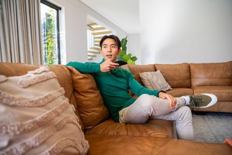 Asian man sitting on brown leather sectional holding remote in modern minimalist living room