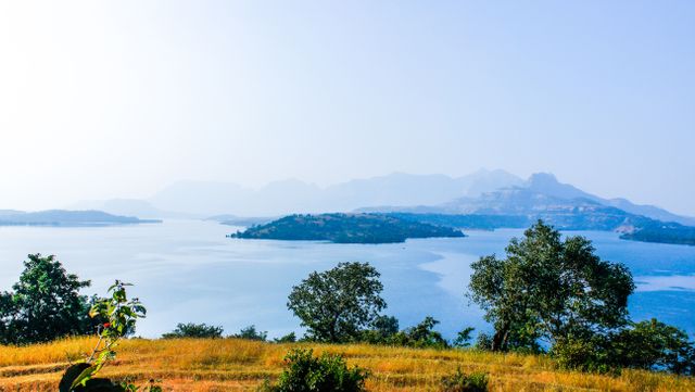 This image captures a serene lake surrounded by lush greenery with distant mountains on the horizon and a clear blue sky above. Ideal for use in travel websites, nature blogs, environmental magazines, or relaxation products ads. The tranquil scenery evokes a calming effect, making it suitable for stress-relief visuals and meditation apps.