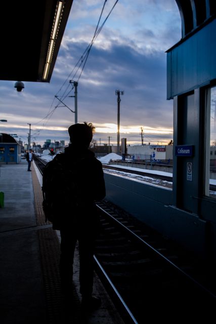 Solitary figure silhouetted against a dramatic sunset sky, waiting on train platform. Suitable for themes of travel, solitude, transportation, urban life. Ideal for blog posts on commuting, travel stories, city living, or transportation-related articles.