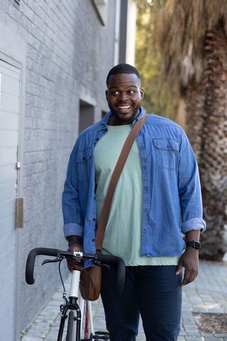 African american cyclist smiling, walking road bike with crossbody bag on urban sidewalk