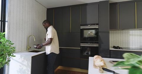 African american man washing dishes in modern dark kitchen with brass faucet and plants