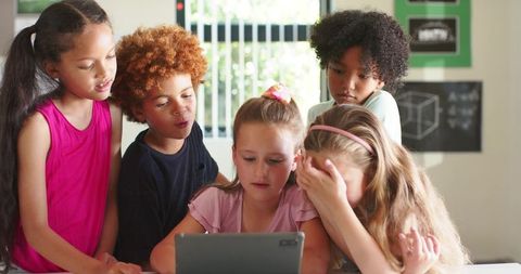 Diverse children learning together around tablet in classroom, collaborating on STEM project
