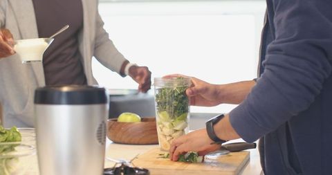 Diverse male friends preparing green smoothie in bright kitchen with blender cup and yogurt