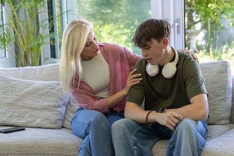 Mother comforting teenage son on sunlit living room couch with headphones and smartphone