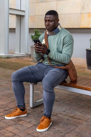 African american man sitting on bench checking smartphone and smartwatch in urban plaza