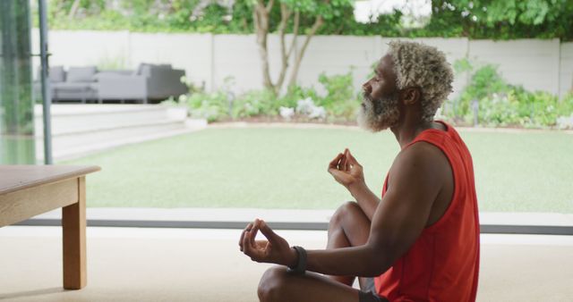 This image captures a senior African American man practicing meditation indoors with an open window that offers a view of a garden. He appears calm and focused, sitting on the floor in a relaxed posture with hands in a standard meditation position, wearing a red tank top. The background includes a well-kept garden and outdoor seating area, which suggests a serene and peaceful environment. This image could be used for articles or marketing materials related to mental health, wellness routines, senior health, mindfulness meditation practices, or promoting a balanced lifestyle.