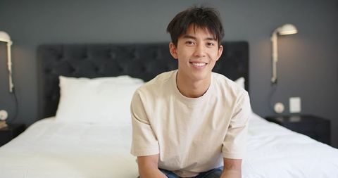 Young man sitting on bed smiling in modern minimal bedroom, cozy casual lifestyle portrait