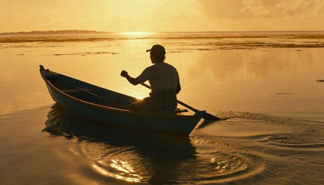 Lone paddler rowing skiff across golden tidal flats at sunset with shimmering water reflection