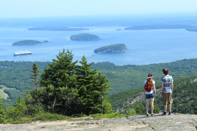 Two people standing on a mountainside, admiring an expansive ocean view with islands in the distance. Ideal for themes related to travel, adventure, exploring nature, hiking, and summertime activities. Perfect for promoting outdoor tourism, hiking gear, or travel locations.