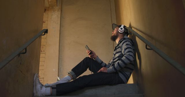Young man in casual clothing sitting alone on stairs listening to music through his headphones while holding a smartphone. Ideal for themes related to urban life, solitude, relaxation, and youthful experiences. This can be used in advertisements for music apps or headphones.