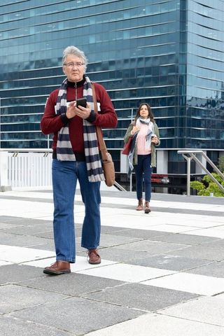 Senior couple walking in modern city plaza, man using smartphone, woman holding coffee