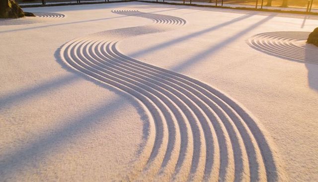 Zen garden raked sand with s-shaped grooves at sunrise, long shadows and boulders
