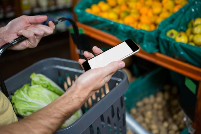 Man holding mobile phone and grocery basket while shopping for vegetables in supermarket. Useful for illustrating modern shopping habits, technology in daily life, grocery shopping, and healthy lifestyle choices.