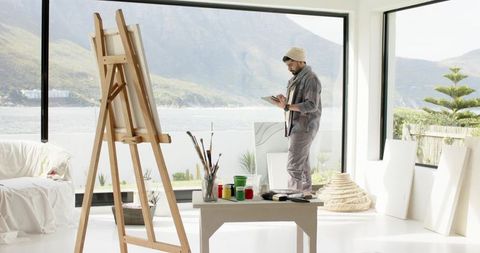 Indian artist sketching on tablet in minimalist waterfront studio with easel and canvases