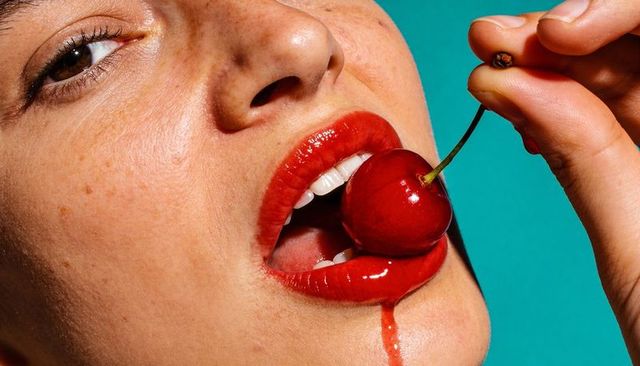 Biting glossy red lips holding ripe cherry with dripping juice and freckled skin on turquoise
