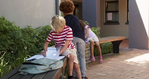 Teenagers studying together on outdoor benches in sunny courtyard with backpacks