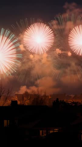 Vertical fireworks display lighting rooftops and bursting pink, green, orange blooms