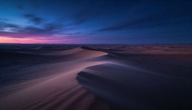 Twilight desert dune ridge with curving crest and wind-blown ripples under deep blue sky