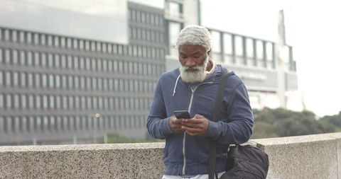 Senior African American man checking smartphone on urban bridge wearing hoodie and duffel